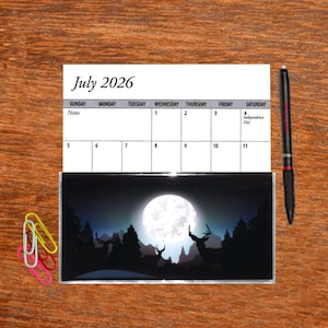 May include: A July 2026 calendar with a black and white image of a full moon and silhouettes of deer in a forest. The text "Independence Day" is written in the calendar for July 4th.