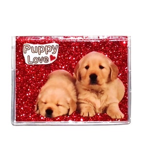 May include: A clear rectangular case with a red glitter background featuring two golden retriever puppies. The text "Puppy Love" is displayed at the top. One puppy is sleeping, and the other is awake.