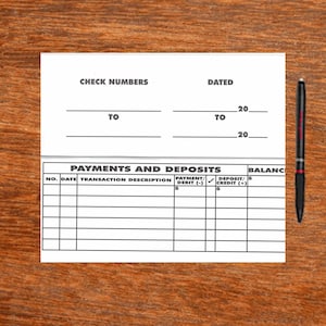 May include: A white checkbook with black text, including "CHECK NUMBERS," "DATED," and sections for payments and deposits. A black and red pen rests on a wooden surface next to the checkbook.