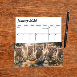 May include: A calendar page for January 2026 featuring a photograph of five kittens in a field of flowers. The calendar has a black pen resting on the right side. The calendar is on a wooden surface.