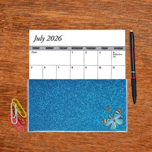 May include: A July 2026 calendar page with a blue glitter notebook with a butterfly design. The calendar page shows the month of July with the 4th of July marked as Independence Day.