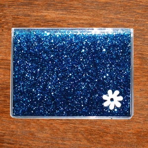 May include: A rectangular, clear case filled with sparkling blue glitter. A white flower is on the lower right corner. The case is on a wooden surface.