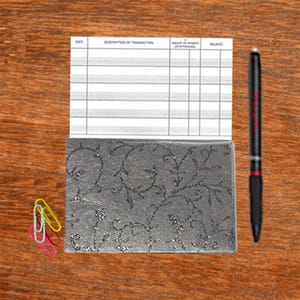 May include: A gray checkbook cover with a floral design. The cover has a silver glitter finish. The checkbook cover is on a wooden surface with a pen and paper clips.