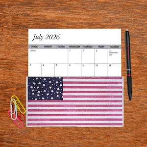 May include: A July 2026 calendar with the 4th of July marked as Independence Day. A small American flag is placed over the calendar.