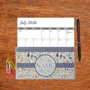 May include: A July 2026 calendar with a floral design and the word "NAME" in a circle. The calendar is on a wooden surface with a pen and paper clips.