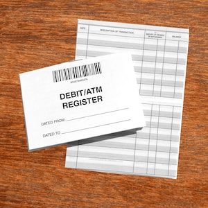 May include: A white Debit/ATM Register booklet with a barcode and the text "DEBIT/ATM REGISTER" is placed on top of a ledger. The ledger has columns for date, description, amount, and balance. The background is a wooden surface.