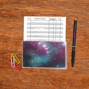 May include: A checkbook cover with a galaxy design, a pen, and colorful paper clips rest on a wooden surface. The checkbook cover features a vibrant nebula of purple, teal, and white. A black pen with red accents is positioned to the right.