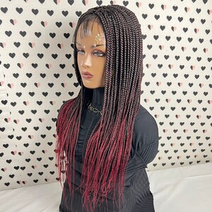 Peut inclure: Une perruque longue tressée ombrée noire et bordeaux avec une dentelle frontale. La perruque est coiffée en style droit et dégagé.