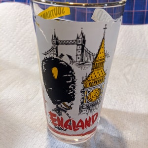 Pode incluir: Um copo de vidro transparente com fundo branco que apresenta uma ilustração em preto e branco do Big Ben, da Ponte da Torre e um soldado britânico. O texto "ENGLAND" está escrito em vermelho na parte inferior do copo.
