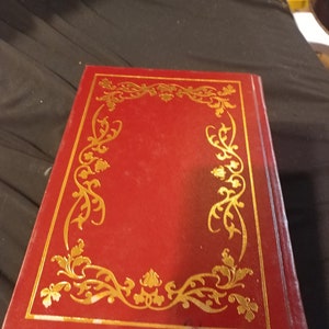 Pode incluir: Um livro encadernado em vermelho com uma borda decorativa dourada na capa.