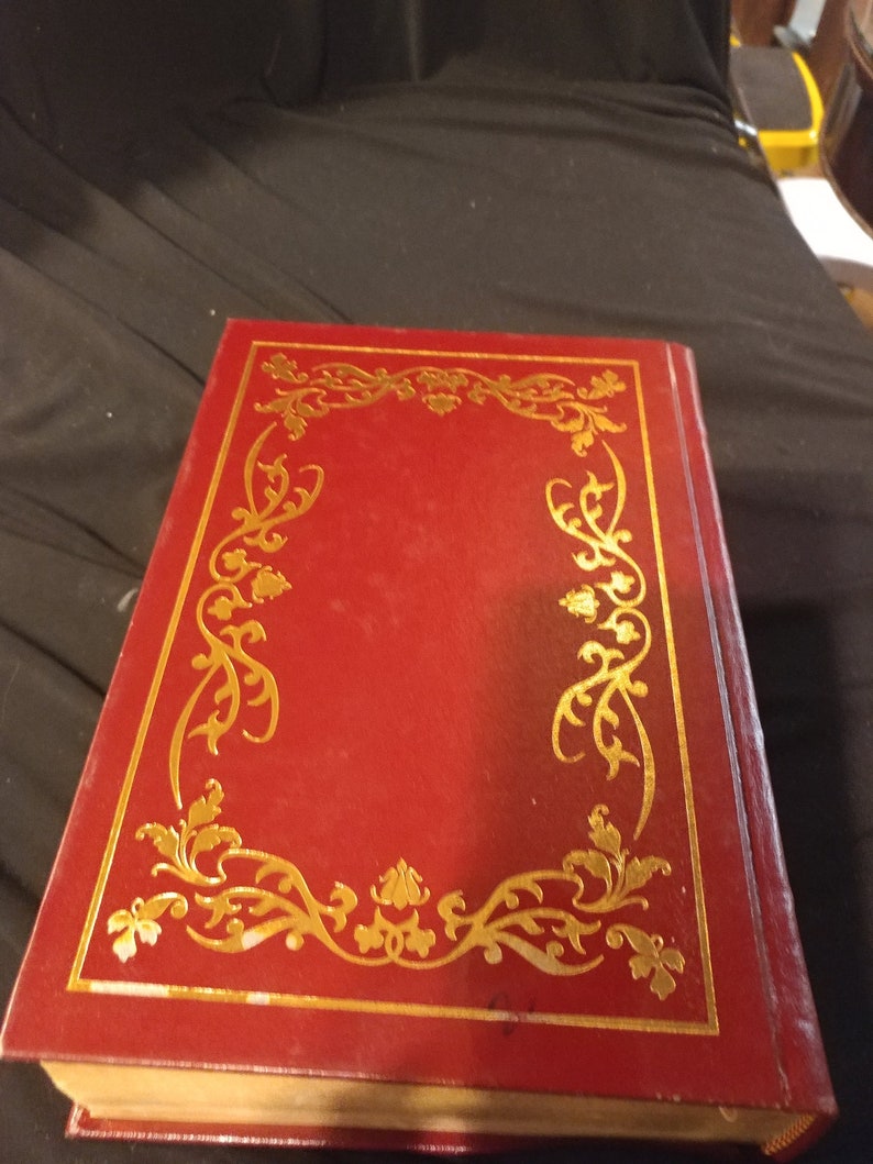 Pode incluir: Um livro encadernado em vermelho com uma borda decorativa dourada na capa.