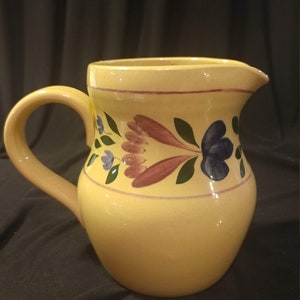 May include: A yellow ceramic pitcher with a floral design in shades of blue, green, and pink. The pitcher has a wide mouth and a handle.