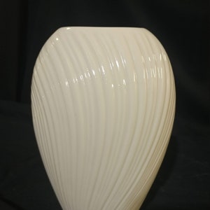 May include: A white ceramic vase with a wavy, ribbed design. The vase is oval shaped and has a smooth, glossy finish.