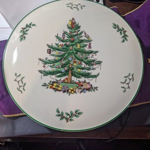 May include: A white ceramic Christmas platter with a green rim features a colorful illustration of a Christmas tree decorated with ornaments, presents, and a bird. The tree is surrounded by holly leaves.