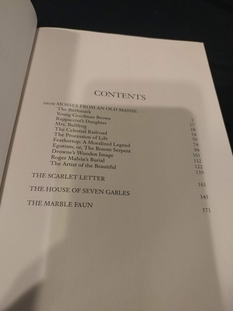 Pode incluir: Um livro intitulado "Mosses from an Old Manse" de Nathaniel Hawthorne. O livro cont&eacute;m uma tabela de conte&uacute;do listando os t&iacute;tulos das hist&oacute;rias e seus n&uacute;meros de p&aacute;gina. As hist&oacute;rias incluem "The Birthmark", "Young Goodman Brown", "Rappaccini's Daughter", "Mrs. Bullfrog", "The Celestial Railroad", "The Procession of Life", "Feathertop: A Moralized Legend", "Egotism; or, The Bosom Serpent", "Drowne's Wooden Image", "Roger Malvin's Burial", "The Artist of the Beautiful", "The Scarlet Letter", "The House of Seven Gables" e "The Marble Faun".
