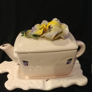 May include: A white ceramic teapot with a square body and a lid decorated with a three-dimensional purple and yellow pansy flower. The teapot sits on a matching white ceramic leaf-shaped tray.