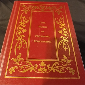 Pode incluir: Um livro encadernado em vermelho com letras douradas na capa. O t&iacute;tulo &eacute; "The Works of Nathaniel Hawthorne".