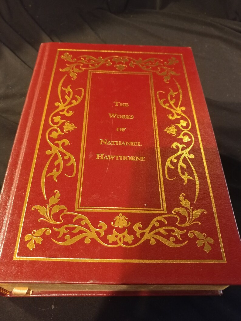 Pode incluir: Um livro encadernado em vermelho com letras douradas na capa. O t&iacute;tulo &eacute; "The Works of Nathaniel Hawthorne".