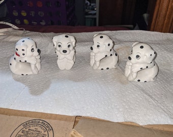 May include: Four ceramic Dalmatian dog napkin rings. The dogs are white with black spots and have their heads resting on their paws.