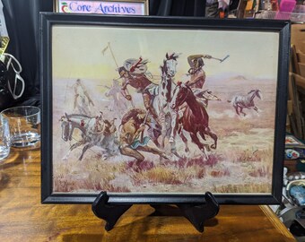 May include: A framed print of a painting depicting a group of Native American warriors on horseback in a battle scene. The warriors are wearing traditional clothing and headdresses, and they are riding horses with colorful blankets. The scene is set in a grassy field with a distant mountain range in the background.