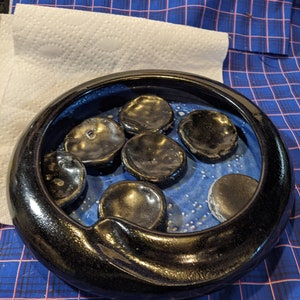May include: A black ceramic bowl with a blue interior and white speckles. The bowl contains eight small, round, black ceramic stones.