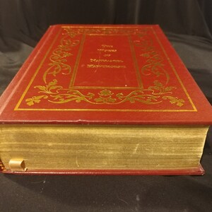 Pode incluir: Um livro encadernado em couro vermelho com detalhes dourados e o t&iacute;tulo "The Writings of Nathaniel Hawthorne" na capa.