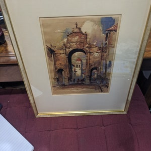 May include: A framed watercolor painting depicting an arched stone gateway in a European city. The painting features a muted color palette of browns, grays, and blues, with a hint of green foliage. The gateway is adorned with intricate details, and the scene is filled with a sense of history and charm.