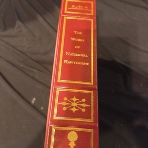 Pode incluir: Um livro encadernado em couro vermelho com letras douradas e detalhes decorativos. O t&iacute;tulo diz "The Works of Nathaniel Hawthorne" e a parte inferior diz "State Street Press".
