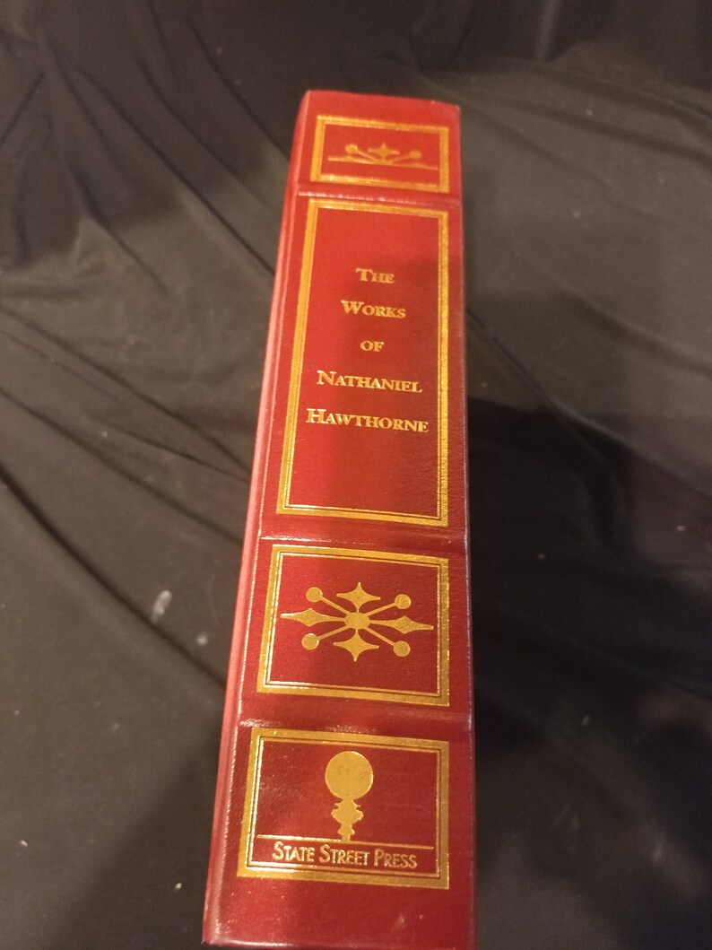 Pode incluir: Um livro encadernado em couro vermelho com letras douradas e detalhes decorativos. O t&iacute;tulo diz "The Works of Nathaniel Hawthorne" e a parte inferior diz "State Street Press".