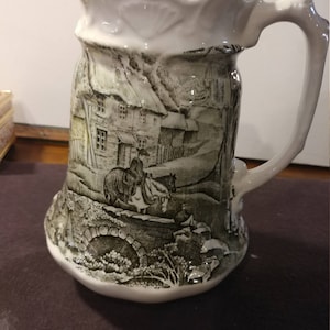 May include: A white ceramic pitcher with a green and white design of a country scene with a house, a bridge, and people on horseback. The pitcher has a ruffled top and a handle.