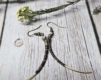 May include: Handmade earrings featuring a curved, bone-like element wrapped in dark wire. The earrings have a hook closure and are set against a rustic wooden background with dried flowers.