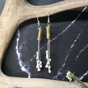 May include: A pair of dangle earrings featuring brass bullet casings attached to silver-toned metal chains. Each earring is adorned with small, white, bone-like charms suspended from the chains, finished with standard silver-toned earring hooks.
