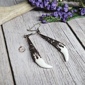 May include: A pair of dangle earrings with a white, tooth-shaped design and intricate bronze filigree. The earrings are set against a rustic wooden background with purple flowers and an antler.