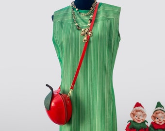 Puede incluir: Un vestido verde sin mangas con rayas blancas, usado en un maniquí. El vestido tiene un bolso rojo con forma de manzana con una correa de cadena dorada. El maniquí lleva un collar dorado con un colgante marrón y dorado.