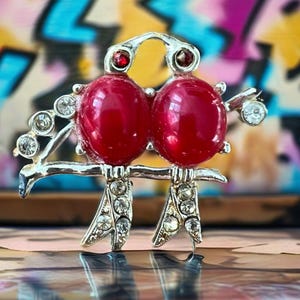 May include: A silver-toned brooch featuring two stylized birds perched on a branch. The birds have large, glossy red bodies and are adorned with clear rhinestones and red accents. The background is a colorful, abstract design.