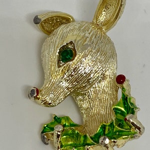 May include: A gold-tone reindeer brooch with a green enamel holly leaf design and a red berry. The reindeer has a green rhinestone eye.