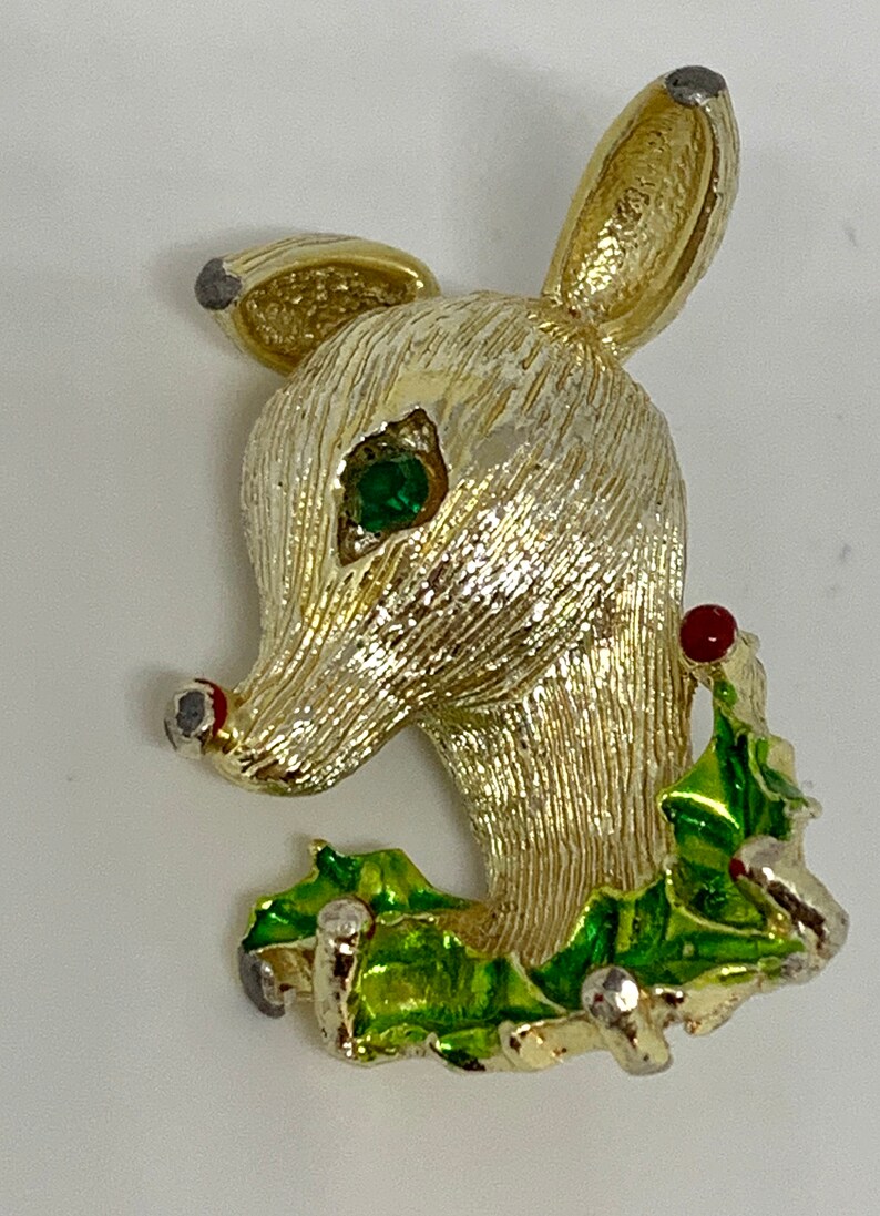 May include: A gold-tone reindeer brooch with a green enamel holly leaf design and a red berry. The reindeer has a green rhinestone eye.