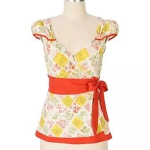 May include: A patterned wrap top with a red sash tied at the waist. The top features a patchwork design with yellow squares and floral accents. Red trim outlines the neckline, sleeves, and hem. The top is displayed on a mannequin.