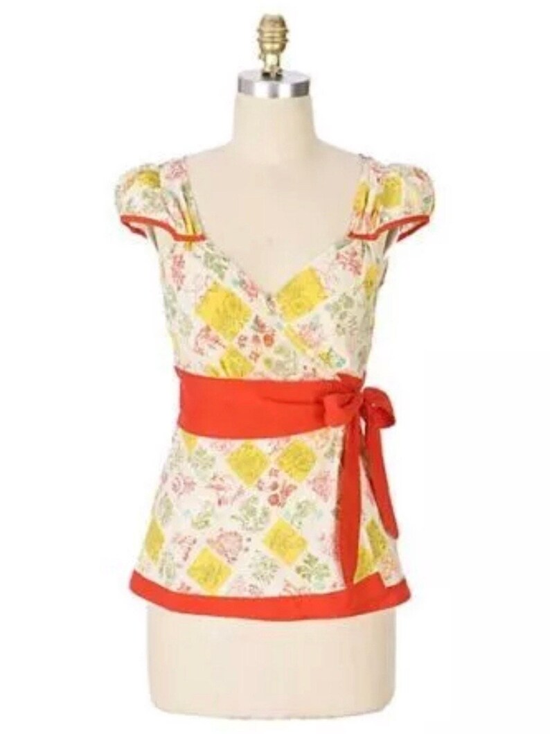 May include: A patterned wrap top with a red sash tied at the waist. The top features a patchwork design with yellow squares and floral accents. Red trim outlines the neckline, sleeves, and hem. The top is displayed on a mannequin.