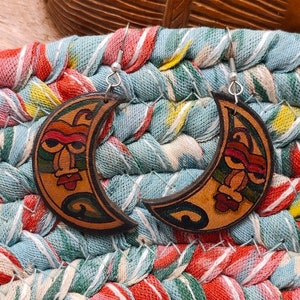 May include: A pair of crescent moon shaped earrings with a hand-painted design. The earrings are made of wood and feature a colorful tribal pattern with red, green, and yellow accents.