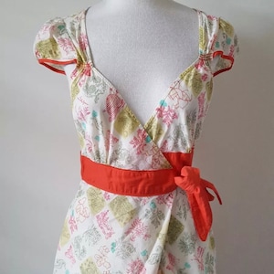 May include: A white wrap top with a floral pattern in pink, green, and blue. The top has short sleeves with orange trim, a V-neck, and an orange waist tie. The top is displayed on a mannequin.