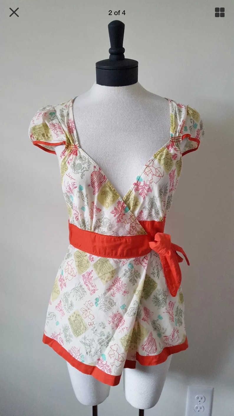 May include: A white wrap top with a floral pattern in pink, green, and blue. The top has short sleeves with orange trim, a V-neck, and an orange waist tie. The top is displayed on a mannequin.