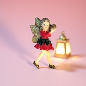May include: A figurine of a fairy with gold glitter wings, a red and black dress, and brown shoes. The fairy is holding a small, round object. A lit lantern is in the background. The background is a solid pink color.