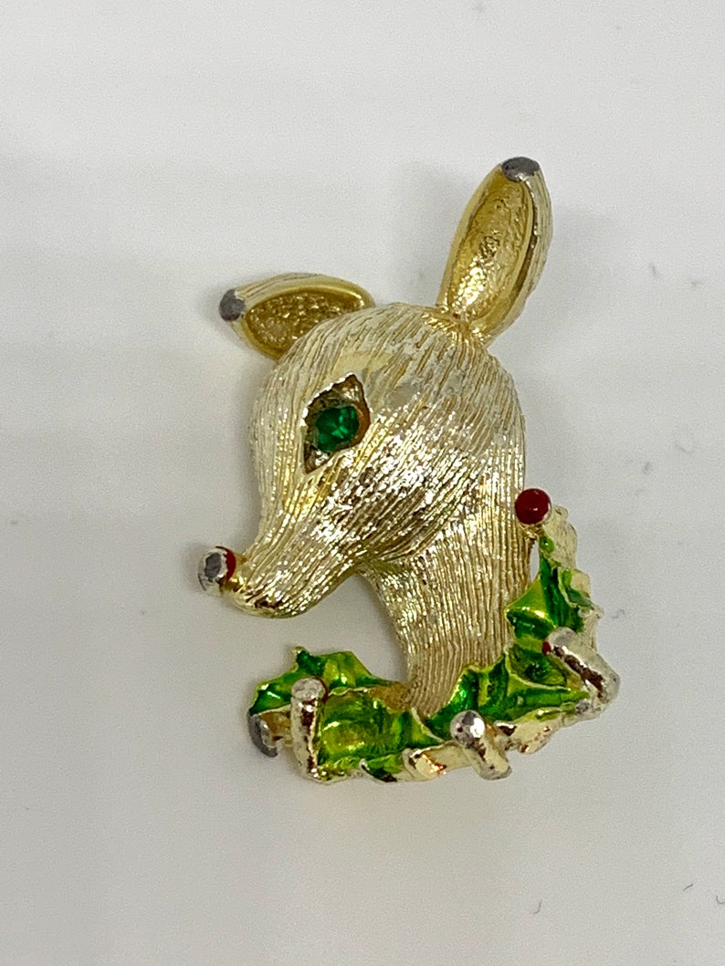 May include: Gold-tone reindeer brooch with green holly leaves and red berries. The reindeer has a green rhinestone eye.