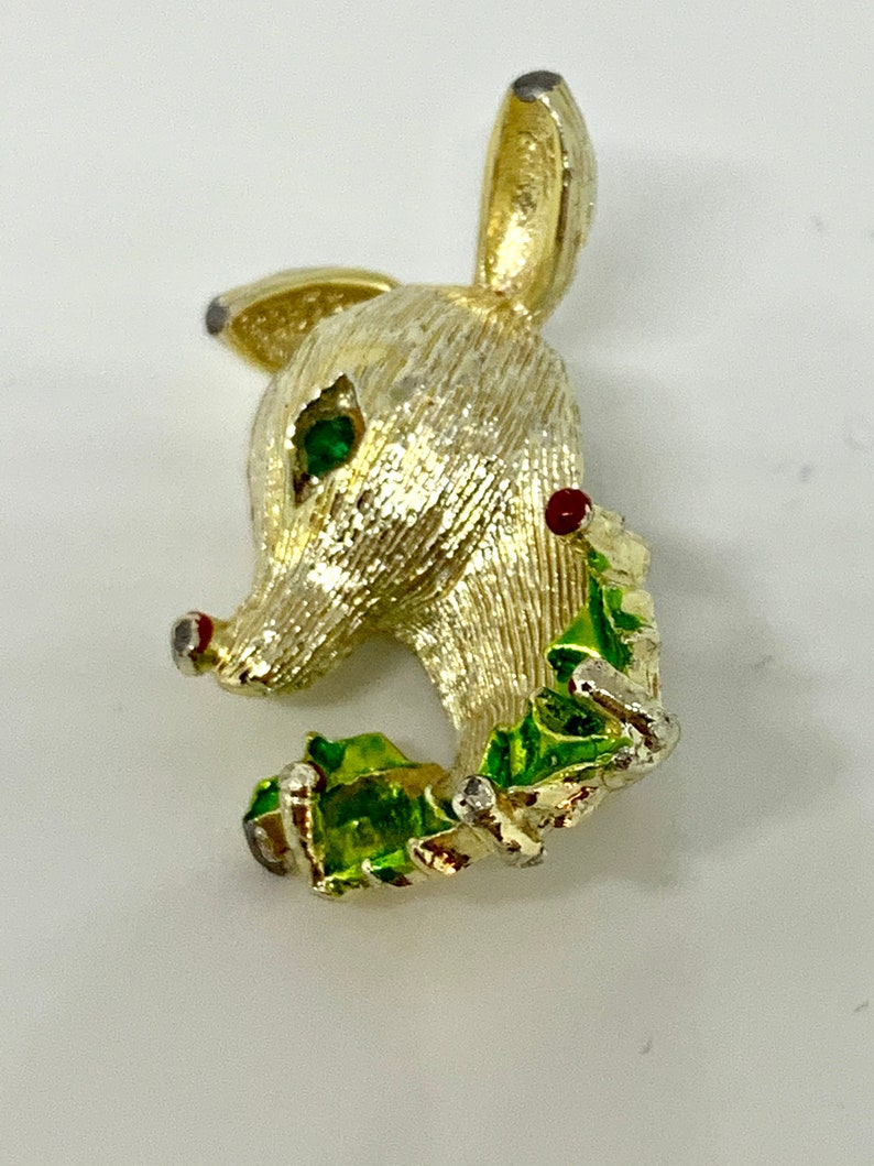 May include: A gold-tone reindeer brooch with green and red enamel accents. The reindeer is holding a sprig of holly in its mouth.