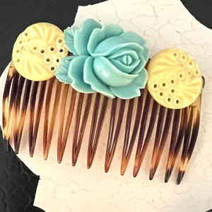 Könnte beinhalten: Vintage Haarkamm mit Schildpattmuster und dekorativen Elementen. Der Kamm hat eine hellblaue Rose in der Mitte, flankiert von zwei gelben, kreisförmigen Akzenten mit einem geschnitzten Muster. Die Zähne des Kamms sind dunkelbraun.