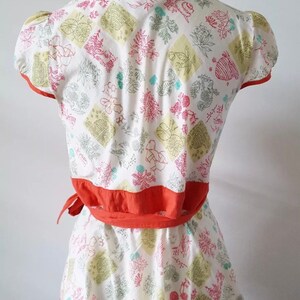 May include: A white two-piece outfit with a floral pattern in pink, green, and yellow. The top has short sleeves and a wrap-around orange belt. The skirt has an orange trim. The outfit is displayed on a mannequin.