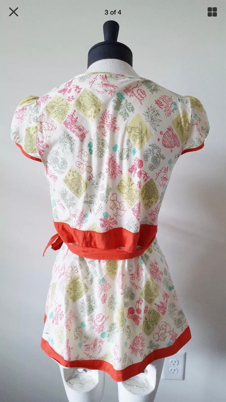 May include: A white two-piece outfit with a floral pattern in pink, green, and yellow. The top has short sleeves and a wrap-around orange belt. The skirt has an orange trim. The outfit is displayed on a mannequin.