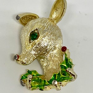 May include: Gold-tone reindeer brooch with a green holly wreath and red berries. The reindeer has a green rhinestone eye.