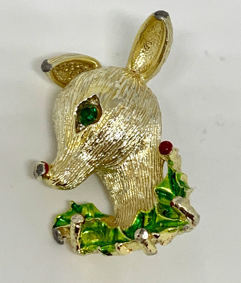 May include: Gold-tone reindeer brooch with a green holly wreath and red berries. The reindeer has a green rhinestone eye.